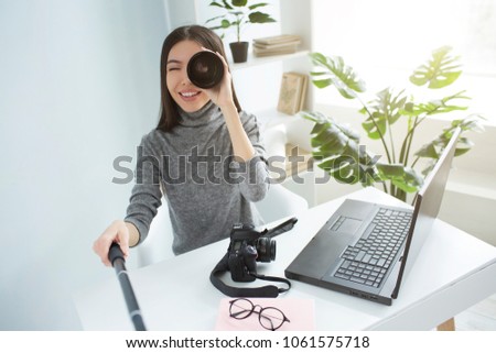 Funny picture of a girl sitting at the table in a big bright room and recording video. She is looking to video camera through the photo camera's lens. Pretty girl is smiling.