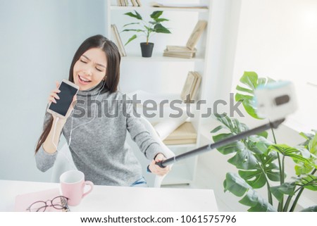 Funny girl pretends she is singing a song and using a phone instead of microphone. She is singing with passion. Also young woman is taking pictures of it using carema on stick for that.
