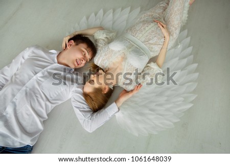 beautiful couple of a man and a pregnant woman in a white lace dress with wings lying on a light floor