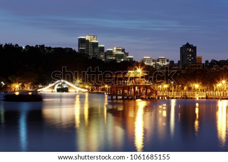 Night scenery of Dahu Community Park in Taipei City, Taiwan, with an arch bridge & an oriental gazebo by the lakeside, residential towers in background under twilight & lights reflected in lake water