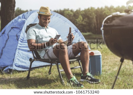Recreation concept. Smiling guy with cellphone and bottle in hands sitting on folding chair. Camp tent on background