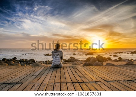 girl sits on a wooden bridge and enjoys the sunset over the ocean. Sunset in Porto, Portugal. Girl tourist looking at the evening sky with orange sun. girl on the beach.