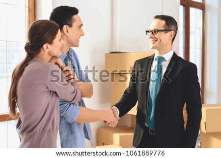 Realtor in suit with family in new apartment with cardboard boxes. Husband and realtor are shaking hands.