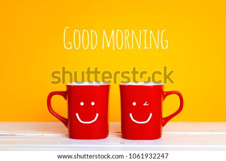 Two red coffee mugs with a smiling faces on a yellow background with the phrase Good morning. Happy coffee mugs.