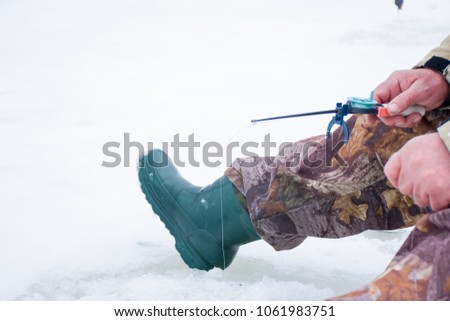 Fisherman sitting on the icy surface of a lake or river and holding a short winter fishing rod for ice fishing. A scene from the winter sport of ice fishing