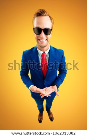 So what?! Portrait of funny young businessman in sunglasses, blue suit and red tie, top angle view shot, over yellow-orange background. Business concept.