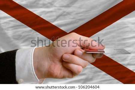 man stretching out credit card to buy goods in front of complete wavy national flag of american state of alabama