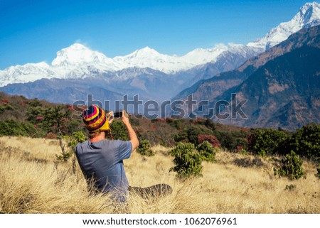 a young tourist man with a hiking backpack and a knitted cap taking pictures of the landscapes and making selfi in the Himalaya mountains. trekking concept in the mountains