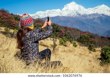 a young tourist woman with a hiking backpack and a knitted hat taking pictures of the landscapes and making selfi in the Himalaya mountains. trekking concept in the mountains