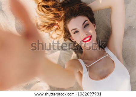 Stunning european lady with red lips making selfie while sunbathing at beach. Overhead portrait of excited girl in white outfit resting on sand.