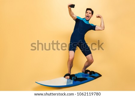 Full length portrait of a strong young man dressed in swimsuit taking a selfie while surfing on a board isolated over yellow background