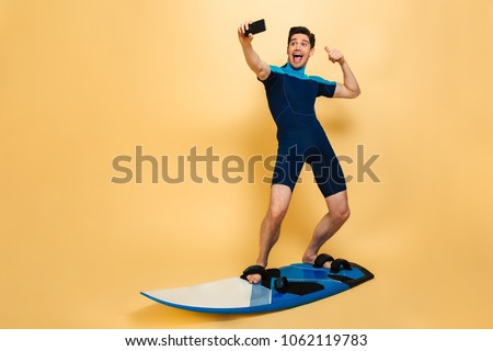 Full length portrait of a joyful young man dressed in swimsuit taking a selfie while surfing on a board isolated over yellow background