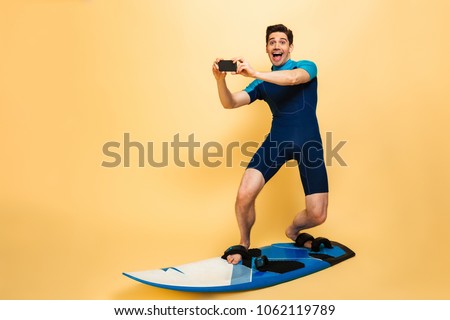 Full length portrait of an excited young man dressed in swimsuit talking a picture with mobile phone while surfing on a board isolated over yellow background