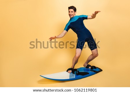 Full length portrait of a smiling young man dressed in swimsuit surfing on a board isolated over yellow background
