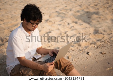 Asian man with his computer on the beach with waves from sea and golden sunlight at evening. working on computer at outdoors while traveling at sea in summer holiday. freelancer work concept.