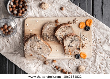 Dessert rye bread with dried apricots, walnuts and prunes lies on a black wooden table with paper. Several pieces are cut off by a sandwich.