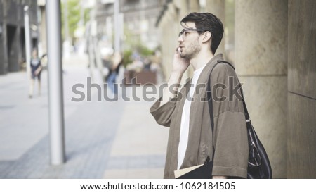 A young man dressed in casual clothes talking on the phone on the street. A young man wearing glasses half-turn