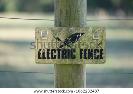 Electric fence sign to keep livestock within the fence, mounted on a wooden post. Blurred background