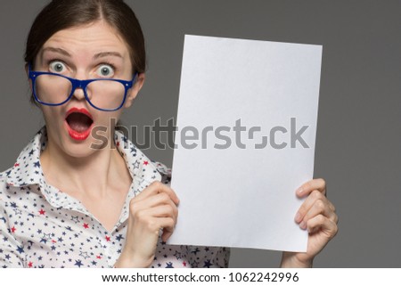 Beautiful brunette girl with glasses holds a white sheet in hands and is surprised on a gray background
