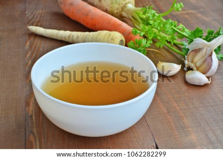 Clear beef broth, bone broth, bouillon in white bowl and vegetables on wooden table 