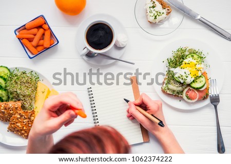 Morning habits of successful people. Day planning and healthy meal. Woman eating carrot and writing in notebook on the served for breakfast white wooden table. Top view, Selective focus Royalty-Free Stock Photo #1062374222