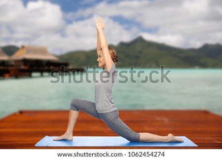 fitness, sport, people and healthy lifestyle concept - happy woman making yoga in low lunge pose on wooden pier over island beach and bungalow background