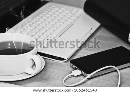 Black and white photo of a cup of hot tea on the table next to the laptop and phone