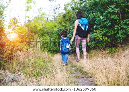 A boy with a backpack is walking through the forest. The child is traveling with his mother. The kid is standing on a trail in the park on the background of trees and holding his mother's hand.