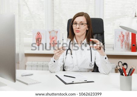 Female doctor sitting at desk with medical documents holds syringe with needle, bottle with medicine in light office in hospital. Woman in medical gown in consulting room. Healthcare medicine concept