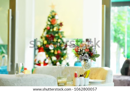 An artificial flower bouquet in a wine glass rests on a dining table and has a Christmas tree ornamented as a background.