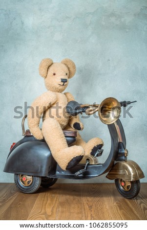 
Teddy Bear sitting on old black retro toy scooter with classic klaxon in front concrete textured wall background. Vintage style filtered photo
