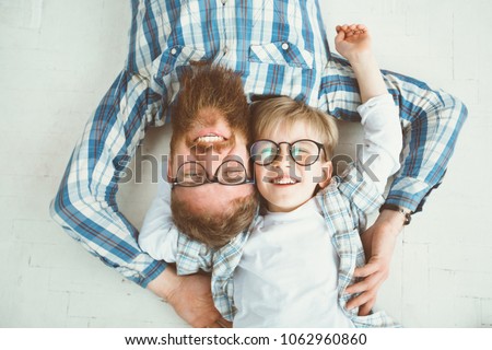 Top view of cute little boy and his handsome young beard dad, both in eyeglasses, smiling while lying with hands behind head on white floor. Royalty-Free Stock Photo #1062960860