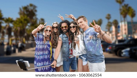 summer holidays, travel and technology concept - smiling young hippie friends taking picture by smartphone on selfie stick and showing peace gesture over venice beach in los angeles background