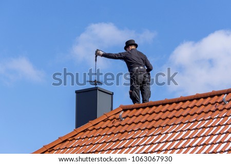 Chimney sweep cleaning a chimney standing on the house roof, lowering equipment down the flue Royalty-Free Stock Photo #1063067930