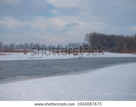 Ice and snow on the riverbank of the Yellowstone River with a deciduous forest and hills in the background and clouds above.