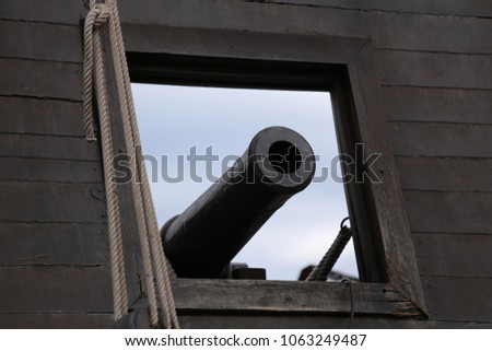 Close up view of an iron canon through an opening in a wooden vessel. Ancient element used during naval warfares. Blue cloudy sky in background. Vintage image of an old historical object. 