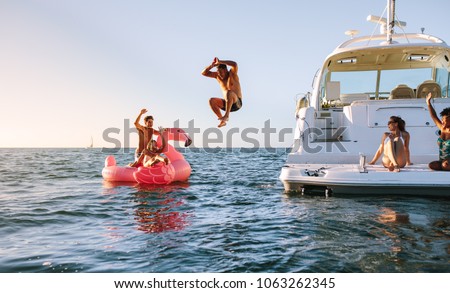 Man diving in the sea with friends sitting on yacht and inflatable toy. Group of friends enjoying a summer day on a inflatable toy and yacht. Royalty-Free Stock Photo #1063262345