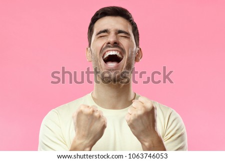 Closeup of emotional man isolated on pink background showing white teeth while screaming with closed eyes, holding hands in gesture of winner, looking extremely happy