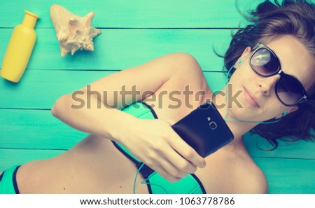 A young girl is sunbathing and listening to music through headphones on a blue wooden background. Accessories for the resort rest on the beach: sunblock, sunglasses, shell. Top view.
