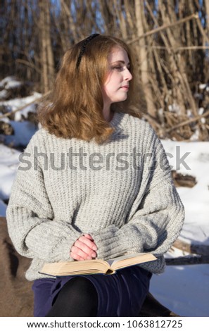 Young beautiful girl in a gray jacket, a purple skirt sits on a rug in a winter forest and reads a book