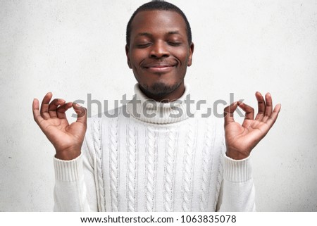 Content dark skinned young male keeps eyes shut and fingers in mudra sign, has calm expression, tries to relax after tired day, wears white sweater, poses against concrete studio background.