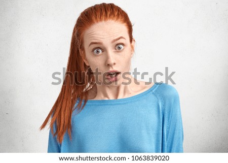 Horizontal shot of amazed beautiful young female looks with stunned expression at camera, feels shock, reacts actively on shocking news, has straight red hair tied in pony tail. Great excitement