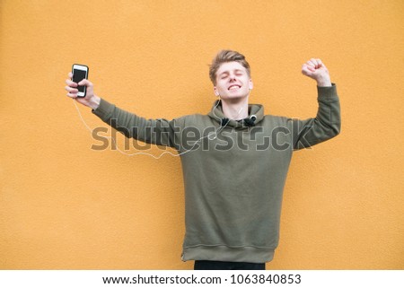 Student enjoys music in headphones while dancing. Happy young man standing in the open air and background of an orange wall and listening to music in headphones