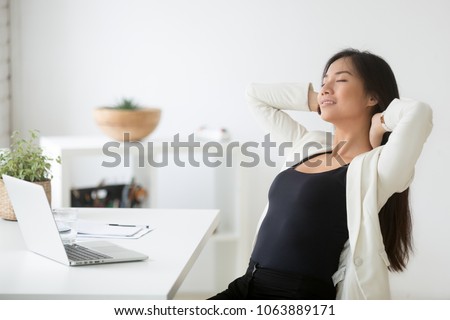 Relaxed happy asian woman enjoying break at workplace, calm smiling korean employee relaxing from computer holding hands behind head breathing air, positive thinking, feeling no stress free at work Royalty-Free Stock Photo #1063889171