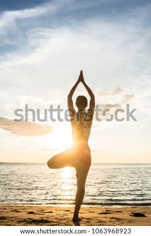 Full length rear view of the silhouette of a woman standing on one leg, while practicing the tree yoga pose on a tranquil beach at sunset during summer vacation in Indonesia