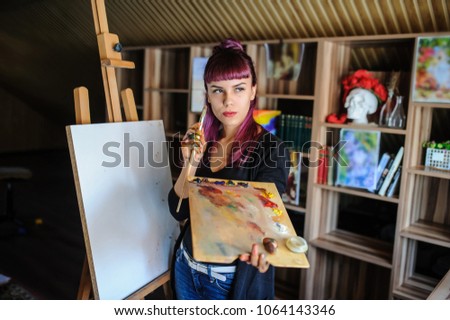 Female artist with purple hair and dirty hands with different paints on them. Holding paintbrush and drawing on white Easel in art studio.