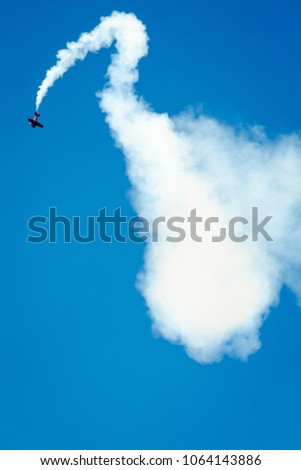 Portrait format airshow photo. Red plane flying upside down releasing white smoke in front of the bright blue sky.