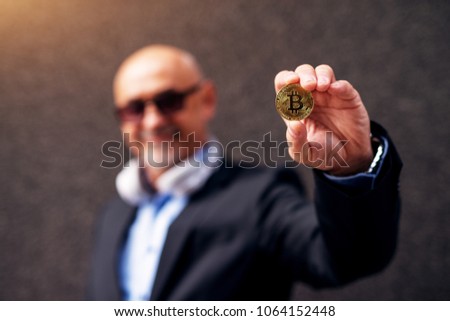 Mature cheerful businessman is extending arm showing a bitcoin to the camera.