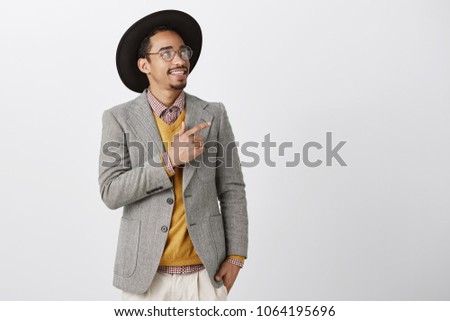 Wealthy businessman seeking new artwork for his house. Good-looking male model in stylish clothes, hat and transparent glasses pointing and looking at upper right corner with interest and amazement