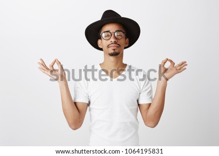 Keep calm and carry on. Studio portrait of relaxed handsome african-american fashion designer in stylish glasses and hat, raising hands with zen gesture, meditating or doing yoga, calming down
